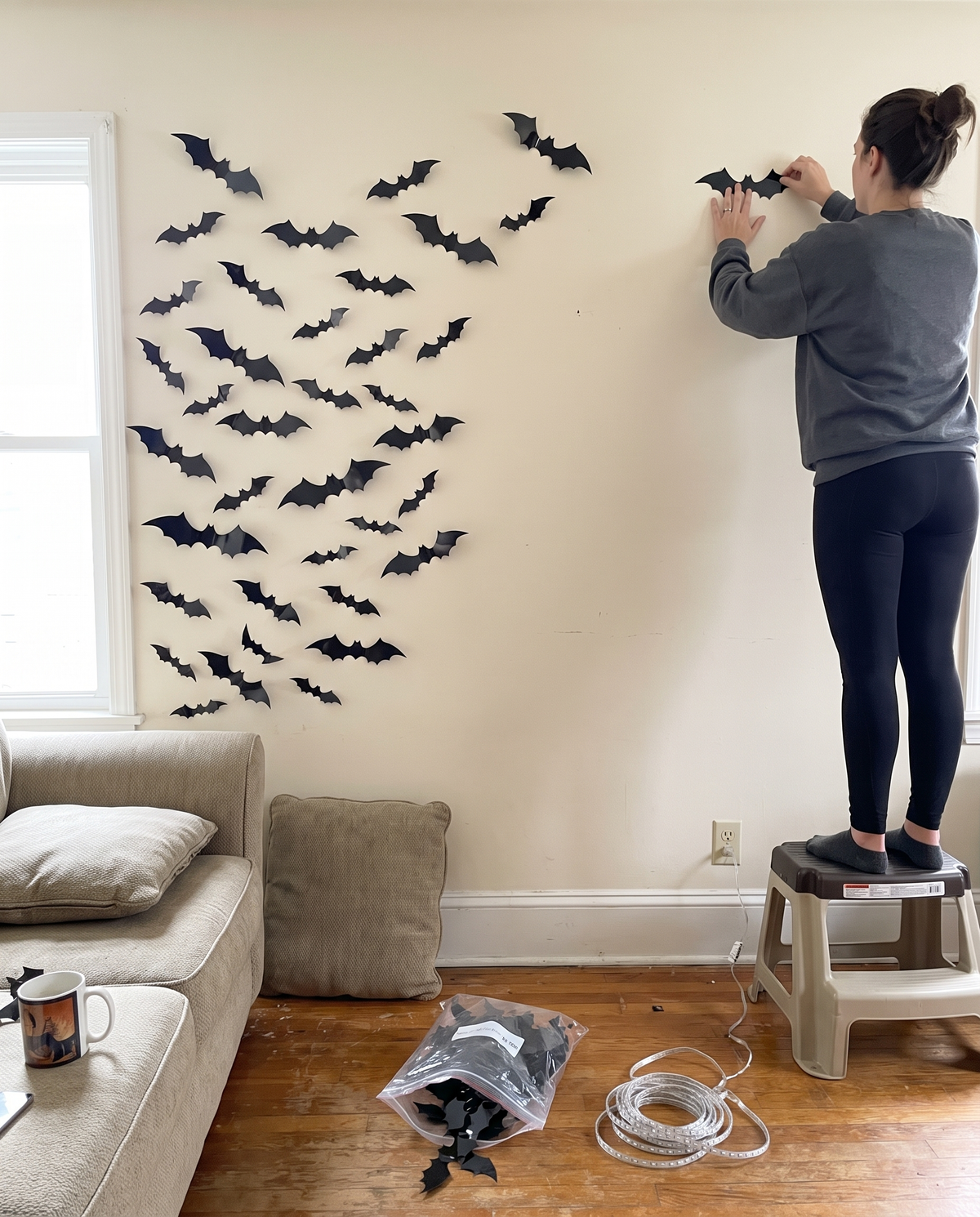 Woman on a step stool sticking black 3D bats to a living room wall, half the swarm done, product bag and LED string on the floor