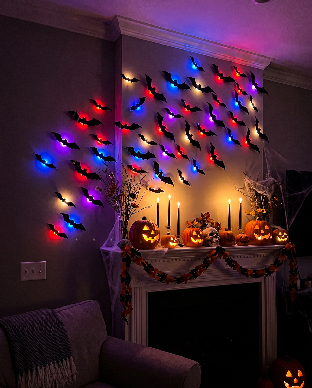 Black 3D bats with colored LED bulbs swarming the wall above a Halloween mantel of lit pumpkins, candles and garland
